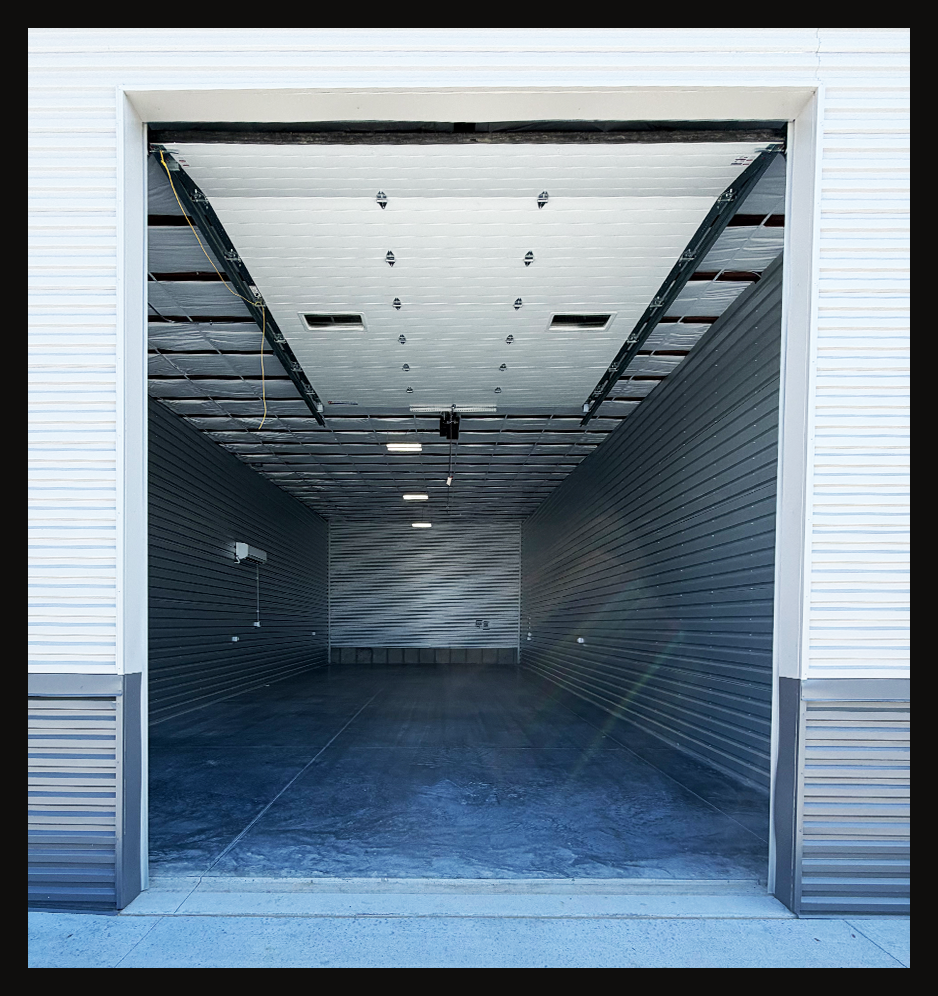 Empty self-storage unit with metal walls and a concrete floor, open roll-up door and ceiling lights.