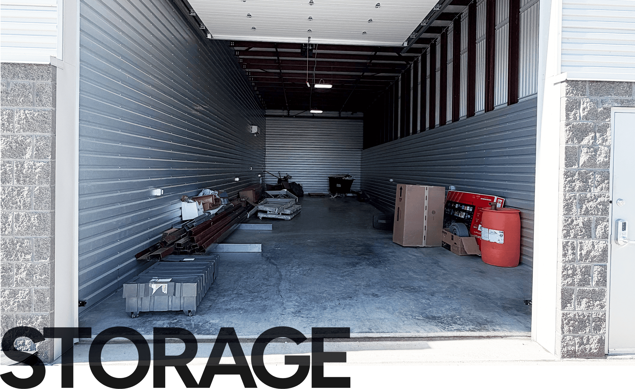 Interior of a storage unit with gray corrugated metal walls, scattered boxes and pallets on the floor, and an orange container to the right.
